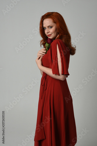 close up portrait of beautiful model with long red hair, wearing an elegant red modern dress. standing pose holding a romantic red rose, isolated figure on studio background.
