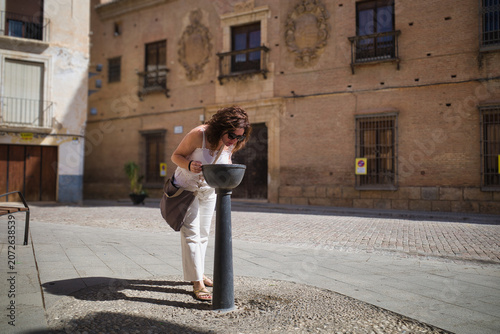 Woman drinks water from public fountain. Historic buildings frame the scene. Sunlight casts long shadows across stone pavement. Her posture suggests quiet, everyday need
