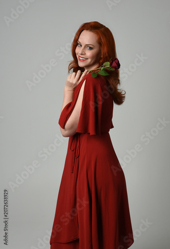 close up portrait of beautiful model with long red hair, wearing an elegant red modern dress. standing pose holding a romantic red rose, isolated figure on studio background.
