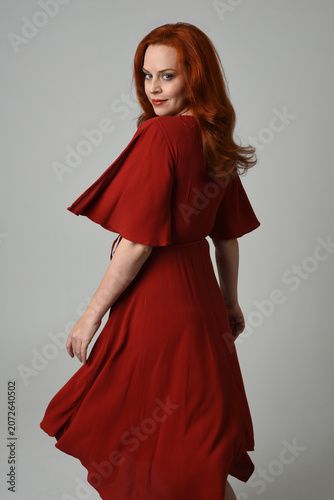 portrait of beautiful model with long red hair, wearing an elegant red vintage dress. standing pose  facing backwards, walking away from camera. isolated silhouette figure on studio background.