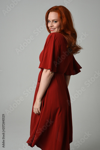 portrait of beautiful model with long red hair, wearing an elegant red vintage dress. standing pose  facing backwards, walking away from camera. isolated silhouette figure on studio background.