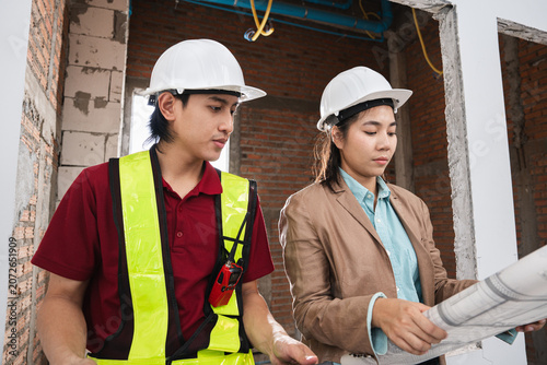 Asian engineer holding paper work and female architect reviewing architectural plans at a renovation or construction site