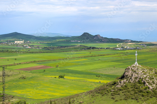 A view of the valley and green meadows, plowed fields. A tall cross on a cliff. Mountains and clouds in the background. Elia, Kakheti, Georgia