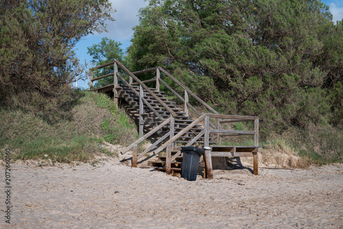 Wooden access stairs leading to a sandy beach in Costa Ballena, Chipiona, Cádiz, Spain, surrounded by coastal vegetation under a partly cloudy sky, representing seaside access and summer tourism.