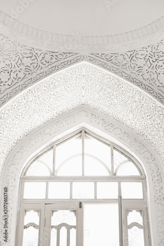 Ornate white plaster carving surrounding a pointed arch window inside Muhammad Amin Khan Madrasah in Khiva. Intricate Islamic arabesques and floral patterns create an ethereal high-key composition.