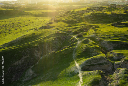 Green hills of Afrasiyab archaeological site near Samarkand at golden hour. Ancient eroded landscape with footpath contrasts with modern city skyline, ideal for travel and heritage publications.