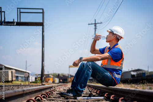 Construction worker drinks water on railroad maintenance site, Railway worker sitting on tracks drinking water to stay hydrated under hot sun, Tired technician taking a break at railway depot