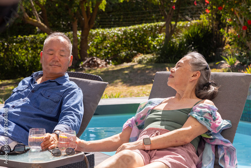 Senior Asian couple reclining poolside holding hands on table with lemonade, in shirt and swimsuit