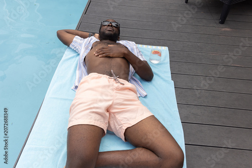 Adult African American man lying and resting on light-blue towel at pool edge, wearing sunglasses