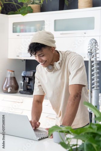 African American man in 20s leaning over laptop in kitchen, smiling with beanie and headphones