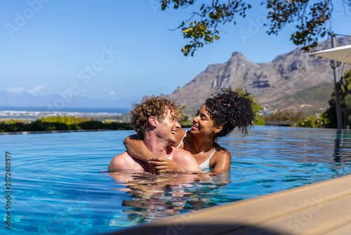 Naklejka Couple embracing and smiling, standing chest-deep in infinity pool by wooden deck, wearing swimwear