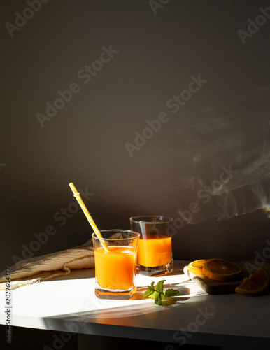 Fresh orange juice in glasses with ice cubes and mint in harsh sunlight. A high-angle studio shot of two glasses filled with vibrant orange juice or citrus smoothie. The scene is illuminated by harsh,