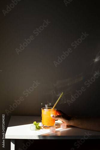 Fresh orange juice in glasses with ice cubes and mint in harsh sunlight. A high-angle studio shot of two glasses filled with vibrant orange juice or citrus smoothie. The scene is illuminated by harsh,