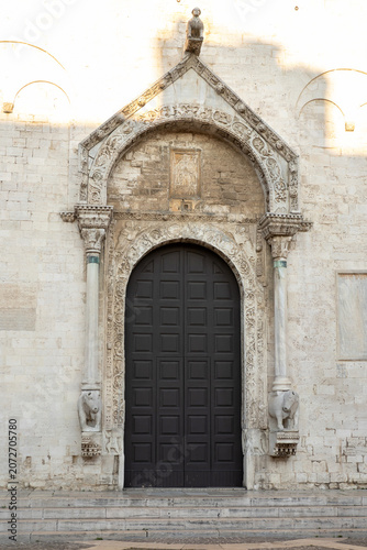 Historic Romanesque cathedral portal in Bari, Italy, featuring an ornate carved stone arch, medieval sculptures, decorative columns, and a monumental wooden door.