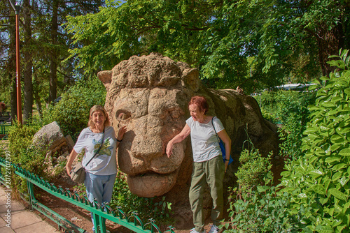 Two female tourists smile happily, posing next to the famous stone sculpture of the Lion of Ifrane in Morocco.