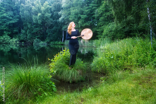 Young redhead woman playing a shamanic frame drum beside a tranquil forest lake, creating a spiritual atmosphere of meditation, rhythm, nature and inner connection