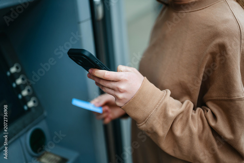 Woman using smartphone and credit card at atm for cash withdrawal