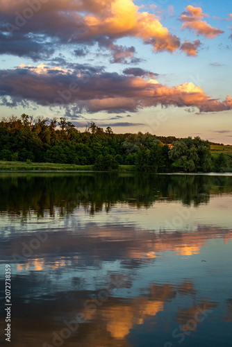 Cuadro en lienzo Warm clouds drift above a green forest and their reflections shimmer on the plac