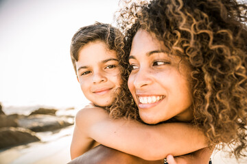 Portrait of a happy mother carrying her son piggyback on the beach