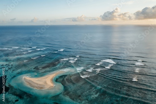 Aerial view of a small sandy island in the middle of the vast blue ocean with gentle waves breaking over shallow coral reefs during sunset