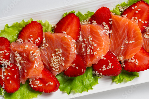 Gourmet salmon slices paired with ripe red strawberry halves on green lettuce leaves sprinkled with sesame seeds served on a white plate representing fusion cuisine, closeup, top view