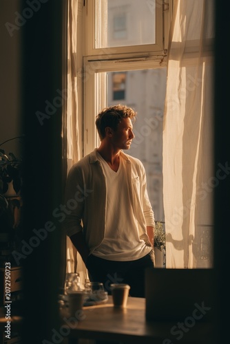 Thoughtful Man Standing by Window in Warm Sunlight at Home