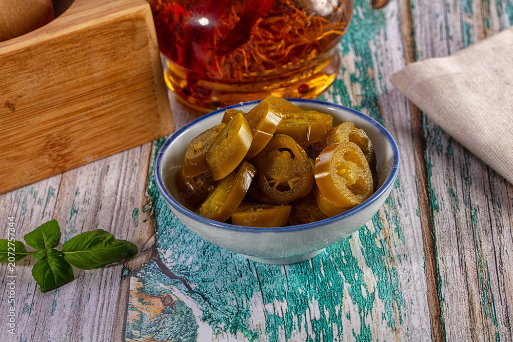 Fototapeta premium Sliced pickled jalapeno peppers in a bowl on a rustic wooden table with chili oil and fresh basil