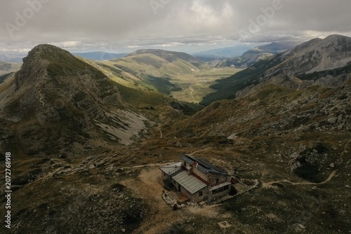 Aerial view of Piani Di Pezza valley with a mountain hut in the foreground, surrounded by rugged mountain slopes and green meadows in Rocca di Mezzo, Abruzzo, Italy.
