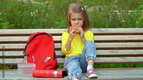 Little school child sits in school yard and eats hamburger. Student girl has breakfast outdoors. Education primary school preschool