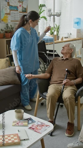Vertical video of young female nurse checking IV drip of senior patient and talking to senior man on sofa. Ideal for nursing home and geriatric care marketing campaigns