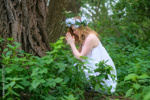Young woman in a white dress kneeling in prayer beside an ancient tree, expressing reverence, spirituality and a deep bond with the living forest