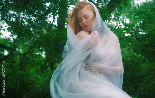 Red haired woman wrapped in translucent fabric stands within a lush forest embracing herself with closed eyes. The portrait evokes vulnerability serenity introspection and quiet renewal