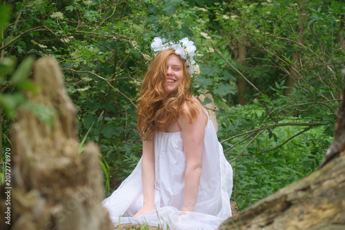 Smiling young ginger woman in a flowing white dress and flower crown in a lush forest, embodying joy, innocence and a magical connection with nature