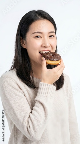 Happy Young Asian Woman in Beige Sweater Eating Chocolate Donut on White Background