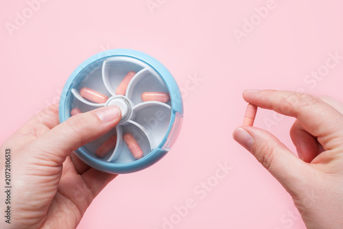 Female hands holding a pill case with pink capsules close-up on a pink background.