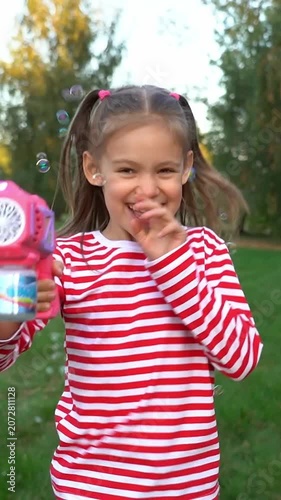 Happy girl run and laugh with bubble gun and soap bubbles in summer park at sunset. Children play in nature. Kids leisure outdoors. Vertical shot