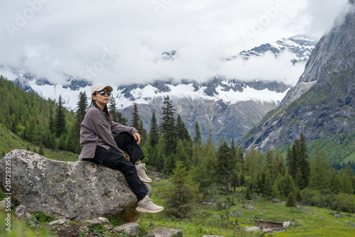 The female tourist sits on a rock, with snow-capped mountains and a forest in the background