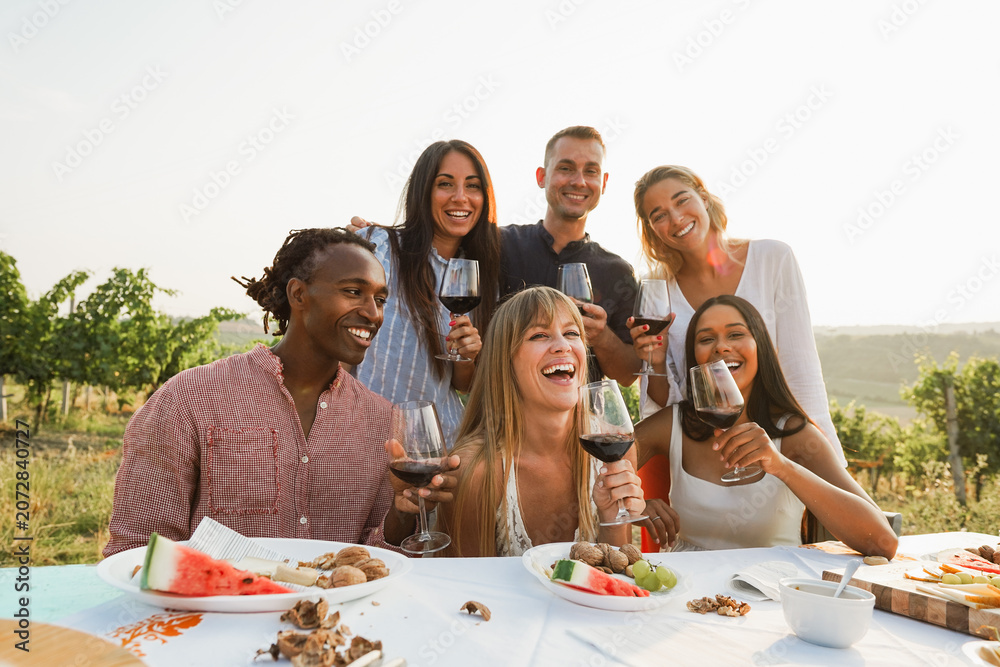 Fototapeta premium Happy adult friends having fun drinking red wine and eating together with vineyard in background - Multiracial people doing party at summer time - Main focus on center girl face - UGC content