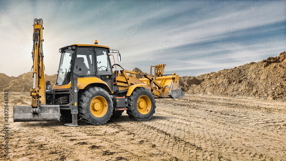 Fototapeta premium An excavator loader operates on a construction site moving soil and materials during daylight. The background shows piles of earth and a clear sky