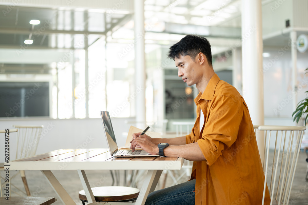 Fototapeta premium Asian man freelancer working on a laptop while sitting at table in comfortable cafe environment.