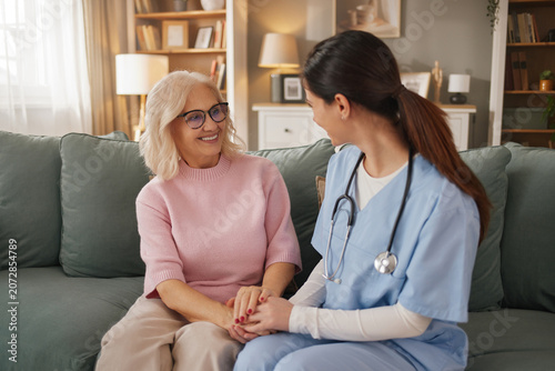 A home health nurse talks with an elderly woman in her living room during a checkup. They are sitting close together, smiling and holding hands.