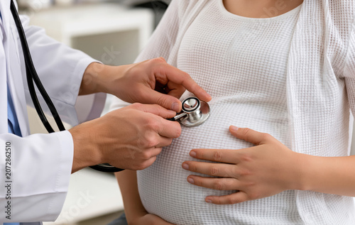 a medical examination is underway with a doctor listening to a pregnant woman's abdomen with a stethoscope. 
