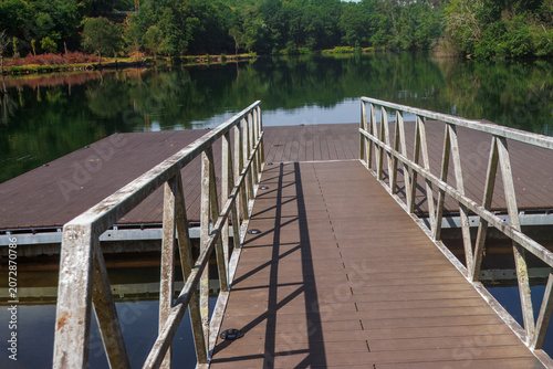 Fotografie Wooden Floating Dock with Metal Railings on a Calm Lake
A perspective shot shows a wooden gangway with weathered metal railings leading onto a floating dock structure on a calm lake