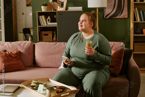 Young plus size Caucasian woman sitting on sofa, holding phone and smoothie while snacking at home, representing mindful routine and self care. Ideal for wellness marketing and lifestyle campaigns