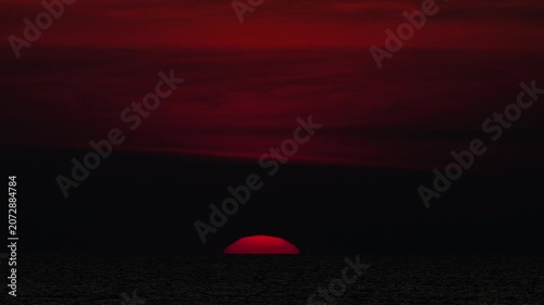 Red sun setting on dark ocean horizon under dramatic crimson sky with clouds. Cinematic wide shot of moody crimson sunset over sea.