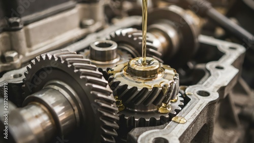Mechanic pouring engine oil onto an open metal gear inside a complex machinery part for lubrication.