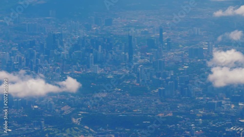 Kuala Lumpur city skyline aerial view from passenger plane window flying above white clouds over dense metropolis on sunny day. Wide angle high altitude slow motion shot.