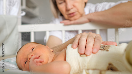 Photos Grandmother's hand gently touching the swaddled newborn baby as it rests in a cr