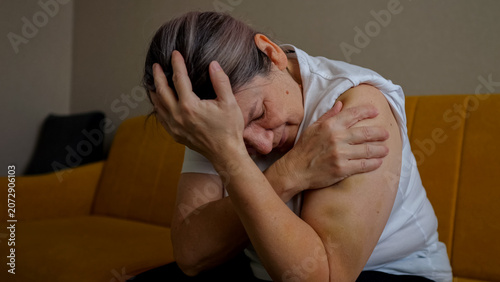 Woman sitting on a sofa, covering her face and holding her arm, expressing emotional pain, sorrow, and being a victim of domestic violence and abuse, highlighting trauma and fear at home