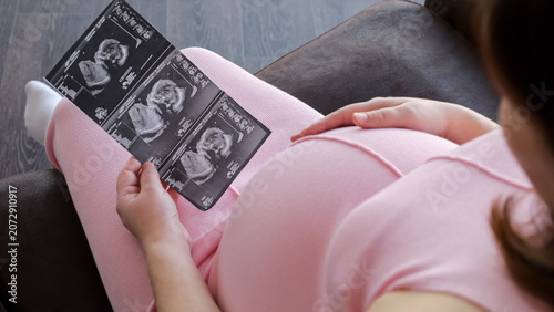 Pregnant woman in pink sitting at home, gently touching her belly with one hand while holding and studying ultrasound printouts, thoughtful and expectant about motherhood
